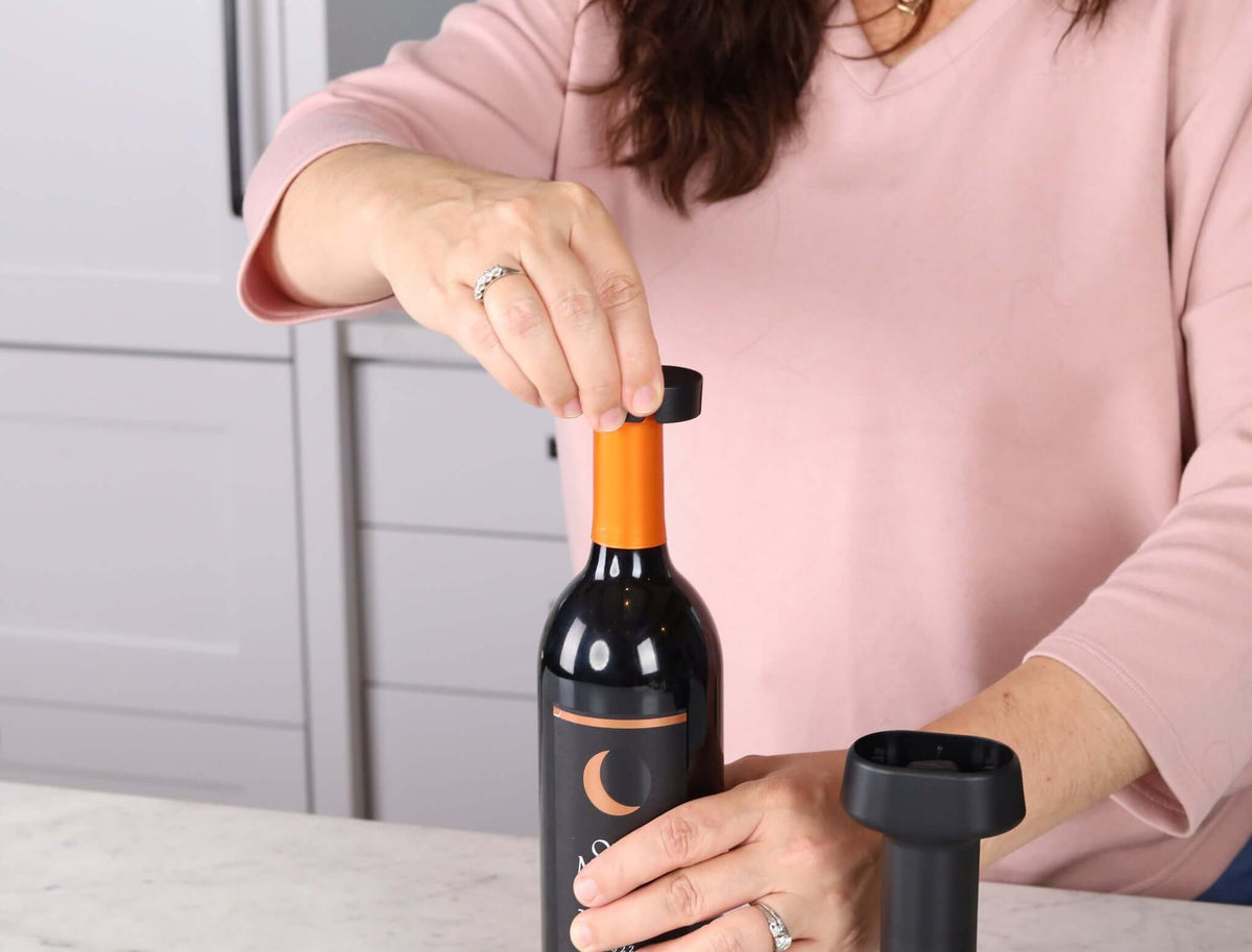Woman using Corkie all-in-one corkscrew opener and wine accessory on a wine bottle in a modern kitchen setting.