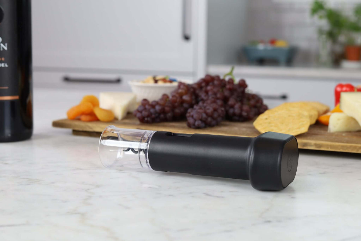 Corkie all-in-one wine tool with corkscrew and pourer on a kitchen counter with grapes and cheese in the background.