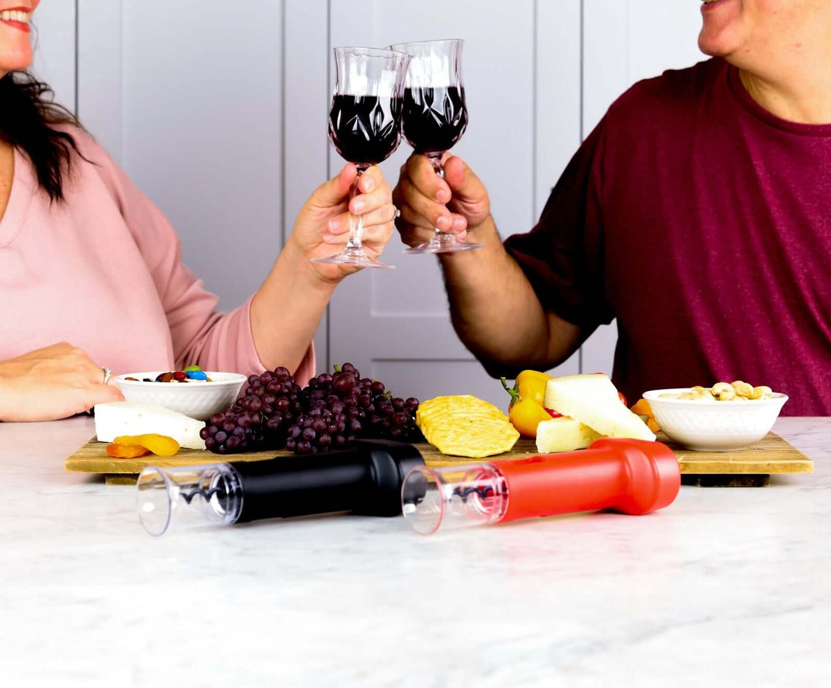 People toasting with wine glasses, showcasing Corkie corkscrew opener sets on a table with assorted cheeses and fruits.