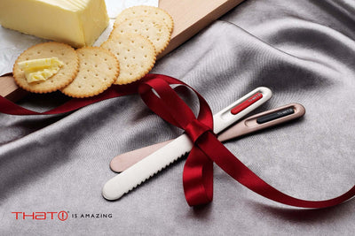 SpreadTHAT! butter knives wrapped in a red ribbon with crackers and butter on a smooth gray surface.